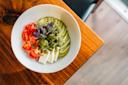 A white bowl on a wooden table contains sliced cucumbers, cherry tomatoes, white cheese, fresh herbs, and a sprinkle of green seasoning arranged neatly.