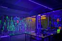 A dark art studio lit by purple and yellow neon lights, with glowing paint splatters on the walls and tables. A Mona Lisa image is drawn in fluorescent paint, with various colorful, glowing decorations hanging from the ceiling.