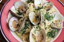 A plate of spaghetti with clams in shells, garnished with chopped parsley and sliced garlic in a light sauce, served in a red-rimmed bowl.