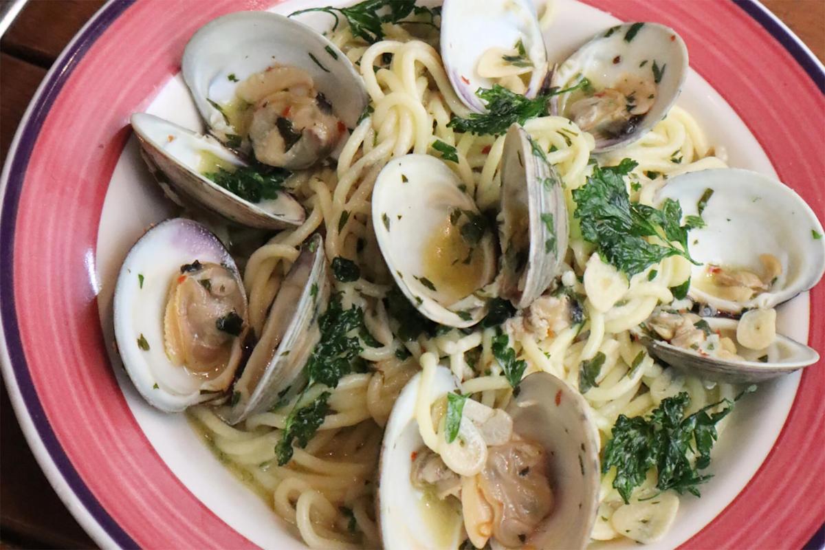 A plate of spaghetti with clams in shells, garnished with chopped parsley and sliced garlic in a light sauce, served in a red-rimmed bowl.