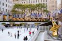 People ice skating at a rink in an urban area. The rink is surrounded by tall buildings with flags on display above it. A golden statue is visible near a water feature on the right.