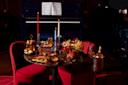 A dimly lit dining setting featuring a round table covered with various elegant dishes, including lobster, meats, and champagne. Two red chairs flank the table, and a piano with sheet music open is in the background. Candles add a warm glow.
