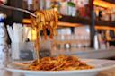 A fork lifts a portion of tagliatelle pasta covered in a rich, meaty sauce from a plate. The background features a blurred view of a bar with shelves of bottles and glassware.