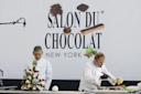 Two chefs in white coats work at a table in front of a “Salon du Chocolat New York” sign. One chef arranges a chocolate sculpture, while the other prepares ingredients with a headset and gloves.