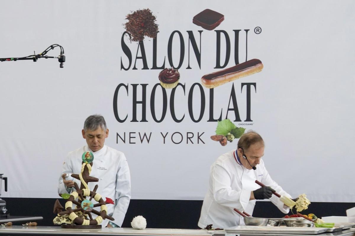 Two chefs in white coats work at a table in front of a “Salon du Chocolat New York” sign. One chef arranges a chocolate sculpture, while the other prepares ingredients with a headset and gloves.