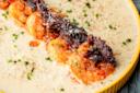 A close-up of creamy shrimp and grits topped with a strip of crispy bacon, garnished with chopped chives and a sprinkle of black pepper, served in a yellow-rimmed bowl.