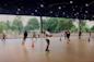 People roller skating under a large covered area with trees visible in the background. The ceiling has decorative lighting, and orange cones are placed around the rink. Skaters of various ages and skill levels are practicing.