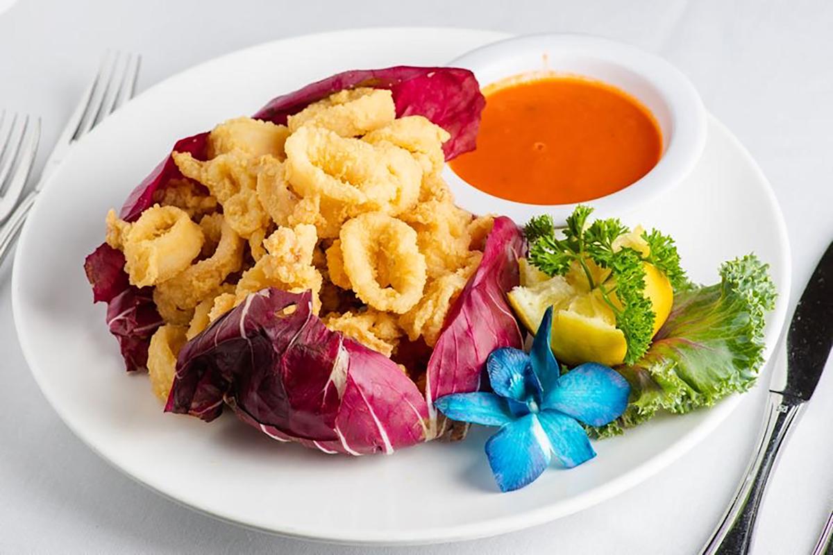A plate of fried calamari served on purple lettuce, with a side of orange dipping sauce, garnished with parsley, a lemon wedge, and a blue edible flower.