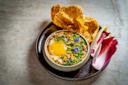 A bowl of savory food topped with a runny egg, herbs, and edible flowers, served with a large crispy cracker and red-chicory leaves on a dark plate against a stone background.