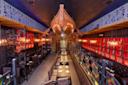 Restaurant interior with a nautical theme, featuring a large wooden boat hanging from the ceiling. Dark leather seating lines both sides of long wooden tables. Warm lighting from hanging fixtures and vibrant red and blue wall art create a cozy atmosphere.