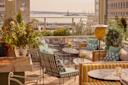 A rooftop terrace with striped chairs, yellow sofa, potted plants, and small tables, overlooking a waterfront with a distant view of the Statue of Liberty and cranes.