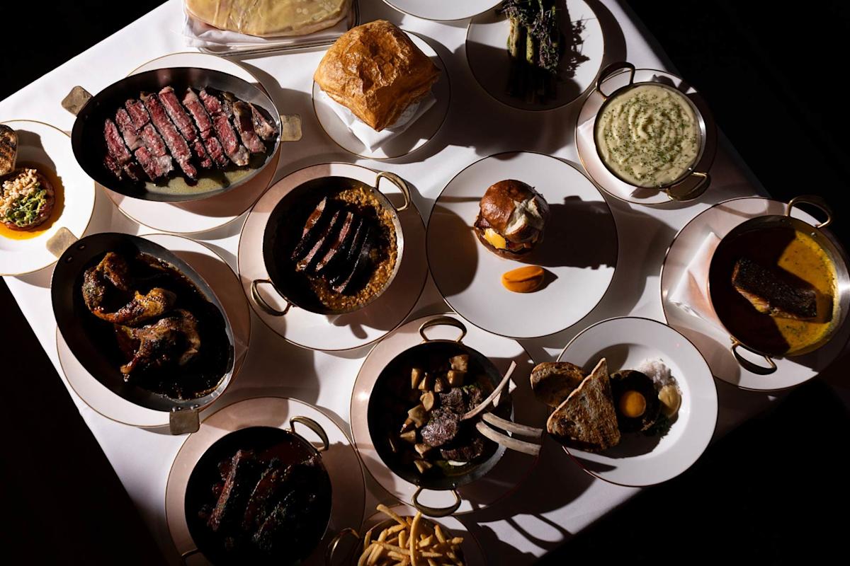 A table set with various plated meats, sauces, mashed potatoes, fries, bread, vegetables, and other sides, all presented in small serving dishes and arranged on a white tablecloth.