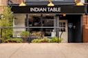 Storefront of a restaurant called "Indian Table" with a black awning and gold lettering. The entrance is accessible with a ramp. There are potted plants and greenery around the doorway and window. Three gold lights are mounted above the awning.