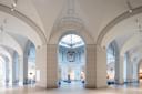 A spacious interior of an art museum with tall, arched ceilings and a central skylight. The floor is tiled, and artworks are displayed along the walls. Large chandeliers hang from the ceiling.