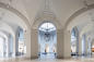 A spacious interior of an art museum with tall, arched ceilings and a central skylight. The floor is tiled, and artworks are displayed along the walls. Large chandeliers hang from the ceiling.