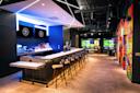 Modern bar with high stools, illuminated blue lighting, and two screens behind the counter. Colorful graffiti art decorates the right wall. In the background, large monitors display golf simulation games. The space is stylish and vibrant.