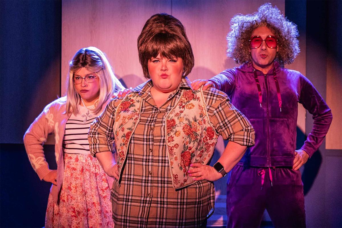 Three people in colorful, retro costumes stand confidently on stage. The one in the center has a large bouffant wig; the others wear glasses, one with a long blonde wig and floral outfit, the other in a purple tracksuit and curly wig.