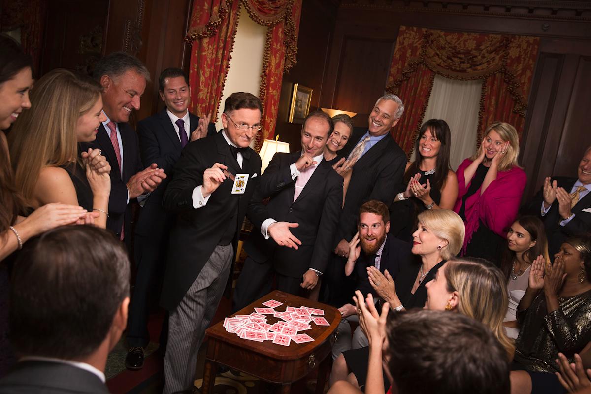 A close-up magician reveals a card to an adult audience in an intimate wood-panelled theatre.