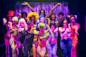 A diverse group of performers in vibrant, colorful costumes pose dramatically on stage with expressive hand gestures, standing in front of a shimmering purple curtain.