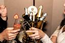 Two people clinking glasses, one with a strawberry cocktail and the other with champagne, near a gold bucket filled with various bottles of sparkling wine.