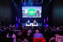 Two people sit on stage in armchairs, speaking to an audience at the Icons of Culture Festival. A large screen behind them displays the event name and a city skyline, with purple and blue stage lighting.