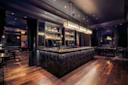 Modern restaurant interior with black and white tiled floor, wooden booths, bar seating, shelves of bottles, industrial ceiling, and colorful graffiti art on the wall in the background.