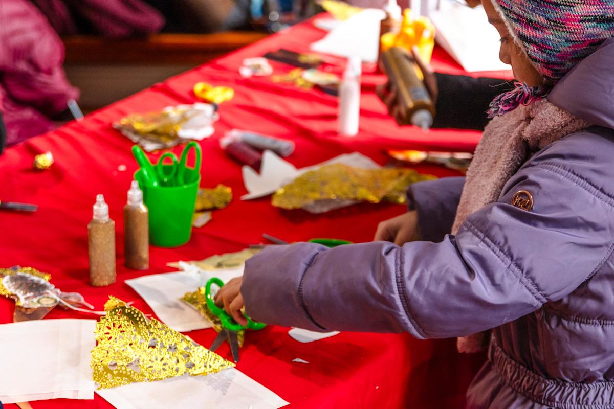 A child wearing a purple jacket and striped hat uses green scissors to cut gold glitter paper at a red table covered in craft supplies, including glue bottles and various materials.