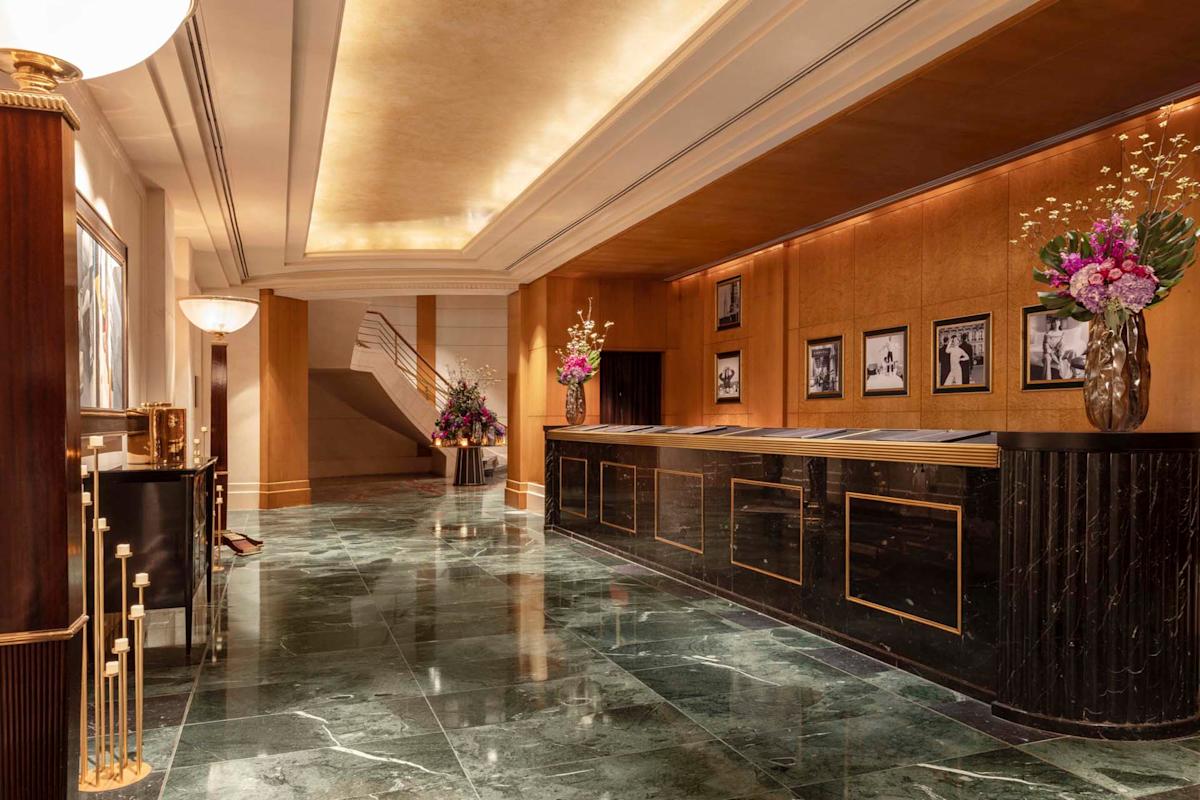 Luxurious hotel lobby with a marble floor, elegant reception desk, and warm lighting. Black and white photos adorn the walls, and flower arrangements add vibrant color. A staircase is visible in the background.