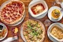 A wooden table set with pepperoni pizza, veggie pizza, lasagna, macaroni and cheese, soup garnished with pumpkin seeds, a cocktail with an orange slice, and a side of bread with dipping sauces.