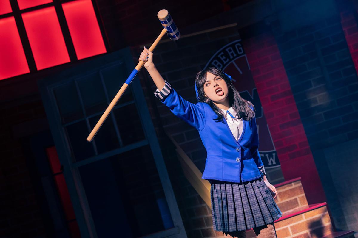 A young woman in a blue blazer and plaid skirt raises a large mallet above her head on stage, with dramatic lighting and a set resembling a school building behind her.