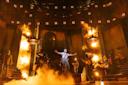 A dramatic stage scene shows a performer singing with arms outstretched amid swirling smoke and orange lighting, surrounded by musicians and actors, evoking an intense and theatrical atmosphere.