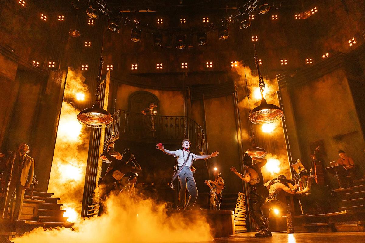 A dramatic stage scene shows a performer singing with arms outstretched amid swirling smoke and orange lighting, surrounded by musicians and actors, evoking an intense and theatrical atmosphere.