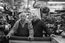 Two older men stand behind a cluttered counter in a shop filled with various items, including an elephant statue and posters. The black-and-white photo captures a cozy, nostalgic atmosphere.