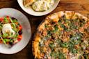 A wood-fired pizza topped with greens, herbs, and cheese sits beside a fresh salad with tomatoes, beets, burrata, and microgreens, and a bowl of creamy ricotta with bread on a rustic wooden table.