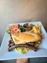 A hand holds a square white plate with a grilled sandwich, sausages, French fries, a side salad with tomato slices, and a small metal cup of dipping sauce.