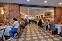 People dining in a cozy restaurant with exposed brick walls and patterned tile flooring. Tables are set with white tablecloths, and soft overhead lighting creates a warm atmosphere. Diners are engaged in conversation, enjoying their meals.