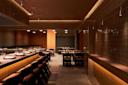 A dimly lit restaurant interior featuring a bar with neatly arranged plates and chairs. The walls and bar are covered in brown tiles, and several tables with chairs are set up in the background. The ambiance is warm and inviting.