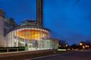 A modern, circular glass building with large windows is illuminated at night. The building features colorful lighting on the ceiling and a cylindrical tower above. A paved road and streetlights are visible in the foreground.
