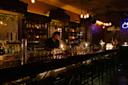 A bartender prepares drinks behind a dimly lit bar with shelves of bottles and glasses, surrounded by dark wood, warm lighting, and empty bar stools in an intimate, cozy atmosphere.