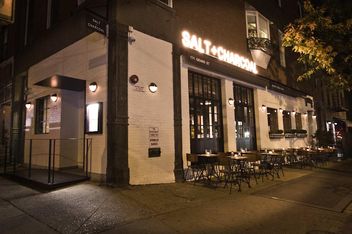 Nighttime view of Salt + Charcoal restaurant exterior, featuring glowing signage, a white brick facade, and outdoor tables with chairs set along the sidewalk, illuminated by wall-mounted lights.