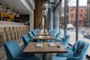 A modern restaurant interior with tables set for dining, featuring plush blue chairs, wooden tables with napkins and utensils, and large floor-to-ceiling windows overlooking a city street.