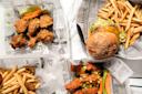 Overhead view of fried chicken drumsticks, a cheeseburger with lettuce, and two servings of French fries, all served in baskets lined with newspaper-style paper.