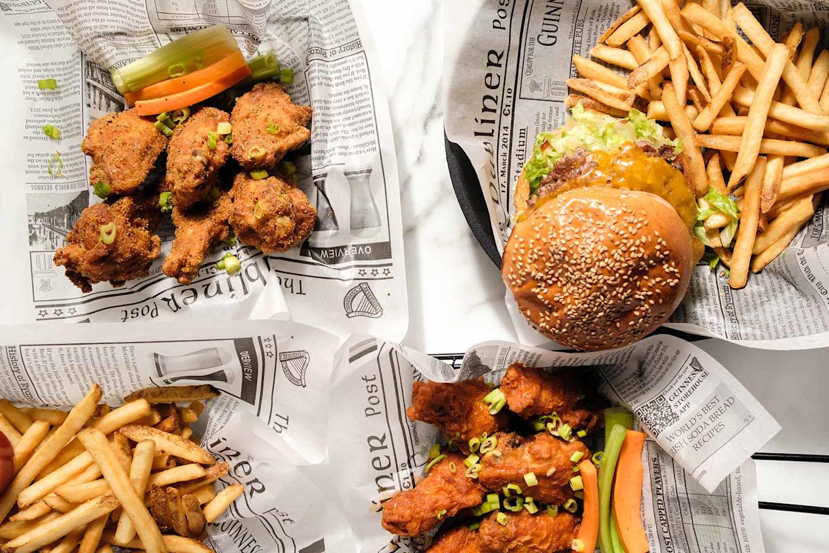 Overhead view of fried chicken drumsticks, a cheeseburger with lettuce, and two servings of French fries, all served in baskets lined with newspaper-style paper.