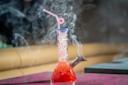 A glass bong filled with ice and red liquid, with smoke swirling around it. A pink bendy straw is inserted at the top, and the bong is set on a maroon surface with a blurred background.
