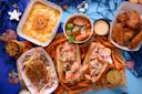 A seafood feast on a blue table with nautical decor. Dishes include lobster rolls, crab cake on coleslaw, fried shrimp, chicken wings, a bowl of soup with sausage and shrimp, and a tray of baked mac and cheese, all surrounded by sweet potato fries.