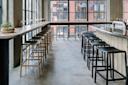Modern, minimal cafe interior with high tables, wooden and black bar stools, potted plant on the table, large windows, and a city street visible outside. The space is filled with natural light.