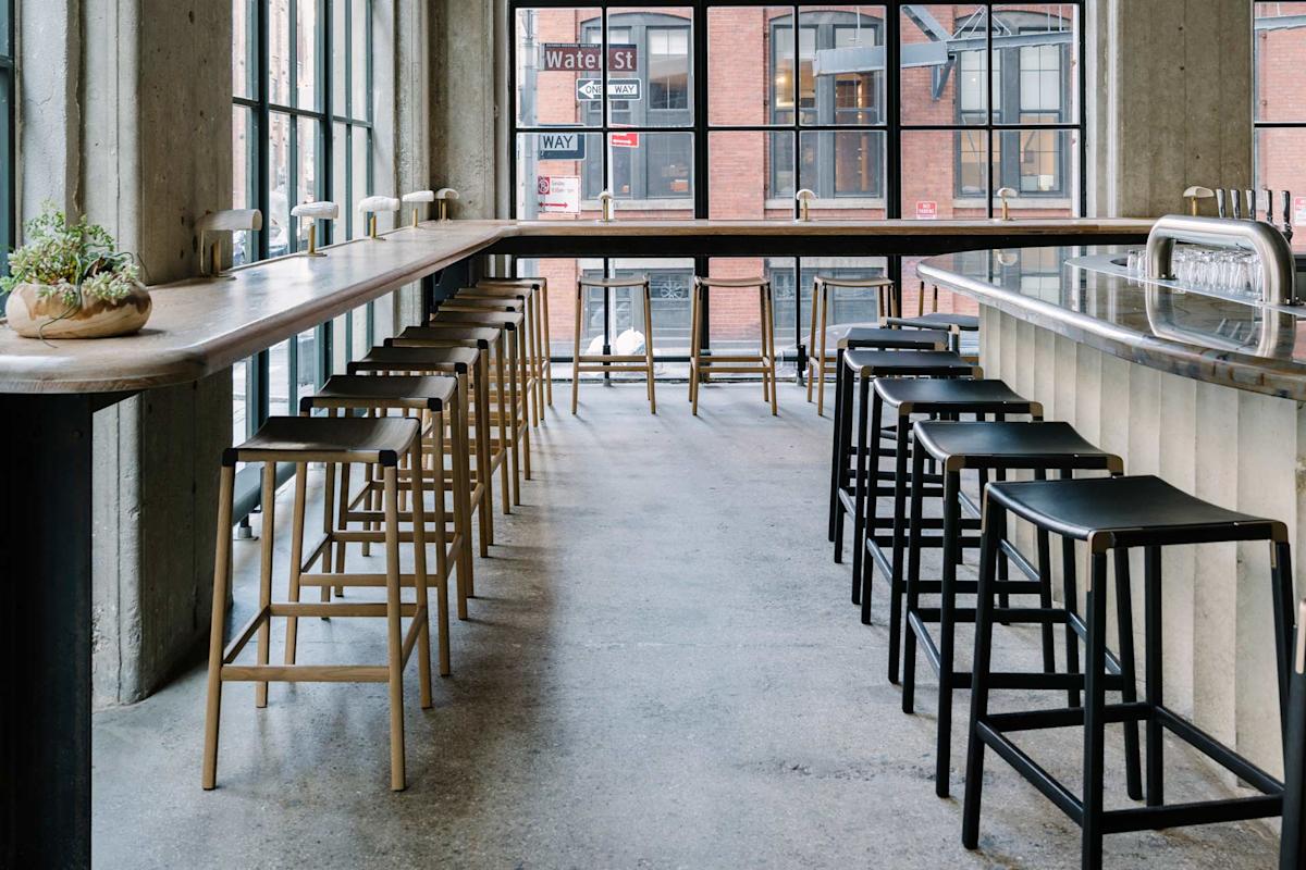 Modern, minimal cafe interior with high tables, wooden and black bar stools, potted plant on the table, large windows, and a city street visible outside. The space is filled with natural light.