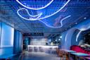 A modern bar with blue neon lights on a grid ceiling, a marble-patterned bar counter, wooden chairs, red velvet booth seating, and a mix of contemporary and reflective decor. The atmosphere is vibrant and stylish.