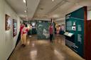 Museum visitors view exhibits about the New Deal, featuring vintage posters, photos, text panels, and a large black-and-white image of a seated woman at the center of the room.