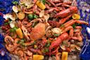 A colorful seafood boil featuring crabs, lobsters, shrimp, clams, and crawfish, garnished with lemon slices and cilantro. Corn on the cob adds a pop of yellow. Layered on a blue fishing net for a nautical theme.